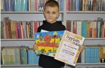 Chłopczyk na tle książek trzymający w dłoniach dyplom oraz prace plastyczną 