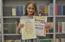 Dziewczynka trzymająca dyplom oraz prace plastyczną stojąca na tle książek w bibliotece