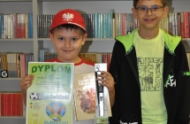 Adam Ćwikliński trzymajacy dyplom i nagrody za udział w konkursie w towrzystwie starszego brata na tle regąłu z ksiazkami