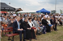 Gosćie dożynkowi i uczestnicy wydarzenia siedż ana łąwkach i stoja w tle