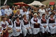 Zespół ludowy Jarzębinki siedzi na ławce, w tle stoją goście dożynkowi