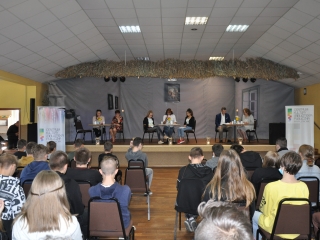 Mlodziez siedzi na krzeslach tylem do aparatu, w tle siedmioro lektorow siedzacycyh przy solikach na scenie