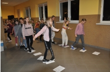 Dzieci w dwóch rzędach przechodzą po kartkach z wyrysowanymi śladami dinozaurów. 