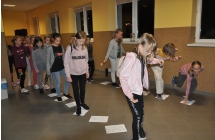 Na podłodze leżą kartki ze śladami dinozaurów, po których przechodzą dzieci ustawione w dóch rzędach