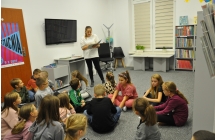 Dzieci siedza na dywanie, w tle bibliotekarka trzymająca w ręce książke, czyta dzieciom