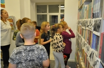 Dzieci przyglądające się ilustracjom dinozaurów zawieszonym na regale z książkami, obok stoi bibliotekarka 