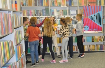 Grupa dzieci stoi przed regałami z ksiażkami, przyglądają sie zawieszonym tam ilustracjom dinozaurów