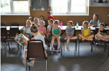 Grupa dzieci siedzi na krzeslach, naprzeciw siedzi dziewczynka i czyta ksiazke