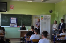 Uczniowie siedza w lawkach, przed nimi siedzi czytajaca kobieta obok ktorej stoi dwoch mezczyzn i kobieta przy banerze CKiBP w Malanowie