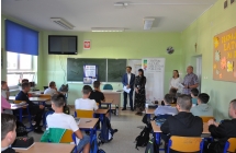 Klasa szkolna, w lawkach siedza uczniowe, przed nimi stoi czworo ludzi, w tle tablica szkolna