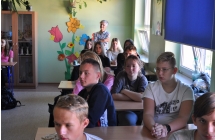 Uczniowie siedza w lawkach w klasie, w tle stoi kobieta