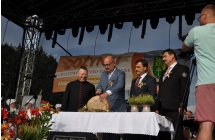 Mezczyzna w towarzystwie kaplana oraz samorzadowcow przekraja chleb dozynkowy