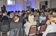 Siedzący ludzie. W oddali scena a na niej zaproszony gość mówiący do mikrofonu 