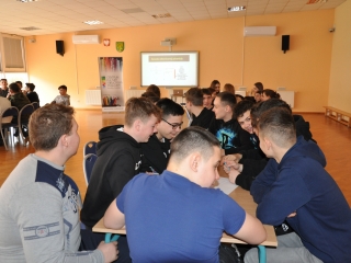 Uczniowie siedza przy stolikach, w tle tablica multimedialna