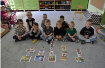 Dziewiecioro dzieci siedzi na szarym dywanie, przed nimi leżą, kolorowe niewielkie obrazki.