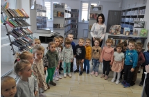 Dziecis toją w półkolu, za nimi stoi kobieta, w tel regały z książkami
