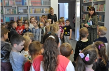 Duza grupa dzieci i dwie kobiety stoją w sali, na ścianach regały z książkami