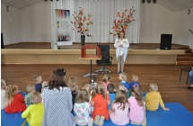 Kobieta stoi przodem do grupki dzieci siedzących na materacach, obok kobiety stoi stolik i krzesło,
