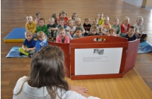 Głowa kobiety obok drewnianej razmy z zadrukowaną karką wewnątrz, w tle dzieci siedzące na materacach