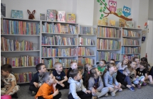 Kilkanaścioro dzieci siedzi na dywanie , za nimi regały z książkami