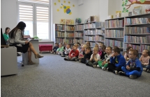 Kobieta siedzi na krześle obok biurka. Czyta książkę dzieciom siedzącym przed nią