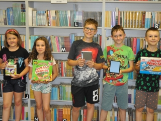 Dwie uśmiechnięte dziewczynki i troje radosnych chłopców. Stoją na tle książek w rękach mając gry planszowe i tablety. 