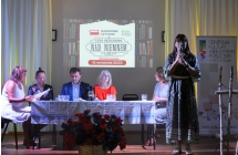 Kobieta mówiąca do mikrofonu na scenie. Obok niej siedzący mężczyzna i cztery kobiety 