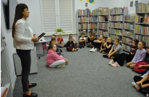 Kobieta mówiąca do dzieci siedzących w bibliotece. W tle książki 
