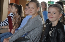 Dwie uśmiechniete dziewczynki siedzące na krzesłach, w tle widoczne inne dzieci 