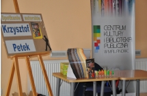 Stolik, na którym lezy stos poukładanych książek. Obok stoi kolorowy roll-up z logo biblioteki oraz plansza z napiszem 