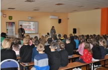 Dzieci siedza na ławkach tyłem do aparatu. Przed nimi stoi mężczyzna i kobieta.