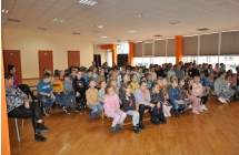Dzieci siedzą na ławkach, przed nimi stoi kobieta i mężczyzna.