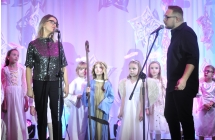 Duet wokalny. Kobieta i mężczyzna śpiewający do mikrofonów. Za nimi grupa dzieci 