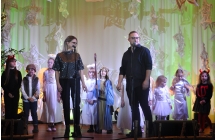 Kobieta i mężczyzna śpiewający do mikrofonów na scenie. Za nimi grupa dzieci. 