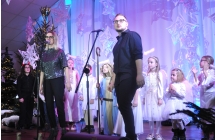 Kobieta i mężczyzna na scenie podczas wykonywania utworu wokalnego. Za nimi grupa dzieci przebranych za kolędników. 