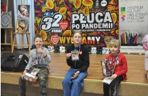 Chłopiec i dwie dziewczynki. Siedzą na scenie. W tle na scenie plakat z logo 32 Finału Wielkiej Orkiestry Świątecznej Pomocy 