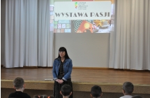 Kobieta mówiąca do dzieci, za nią wyświetla się logo - Pierwszej Wystawy Pasji 