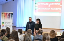 Mężczyzna wraz kobietą. Przed nimi siedzący uczniowie. Spotkanie autorskie.