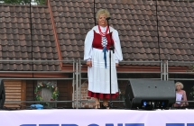 Kobieta ubrana w ludowy strój wykonuje utwór wokalny na scenie 
