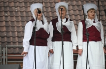 Trzy kobiety w ludowych strojach śpiewające na scenie do mikrofonu 