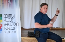 Mężczyzna siedzący na scenie przy banerze z napisem Centrum Kultury i Biblioteka Publiczna w Malanowie 