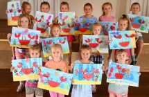 Grupa dzieci stojących przy scenie i prezentujących prace plastyczne 