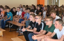 Grupa kilkudziesięciu dzieci siedzących na ławkach i słuchających czytających podczas narodowego czytania 