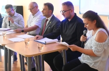 Czworo mężczyzn i kobieta siedzi przy stole. Kobieta czyta do mikrofonu. 