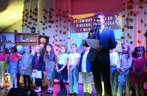 Mężczyzna mówiący do mikrofonu. Za nim grupa kilkunastu dzieci 