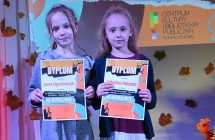 Dwie dziewczynki stojące na scenie. Obydwie trzymają w dłoniach dyplomy