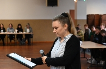 Kobieta trzymająca mikrofon. Stoi na sali w której jest kilkadziesiąt osób 