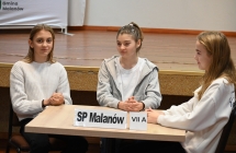 Trzy dziewczynki siedzące przy stole. Przed nimi plakietka z napisem SP MALANÓW