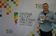 Uśmiechnięty mężczyzna w koszuli trzymający książkę na tle ścianki z napisem - centrum kultury i biblioteka publiczna w malanowie 