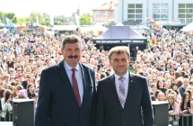 Dwoje elegancko ubranych mężczyzn stojących na scenie. Za nimi kilkutysięczny tłum ludzi. 